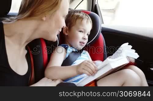 Mother talking to son while they traveling by car. Boy holding open book sitting in child safety seat