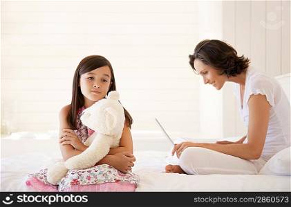 Mother using laptop daughter cuddling teddy sitting on bed