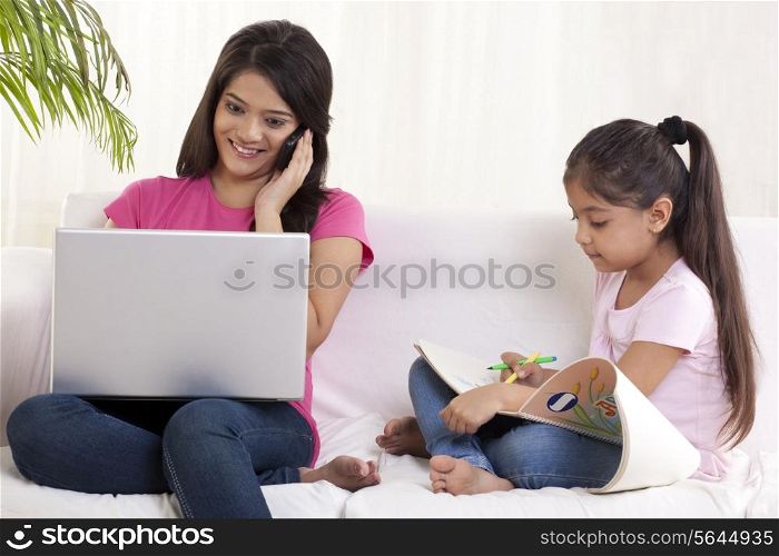 Mother using laptop while talking on phone with daughter sitting besides her and drawing