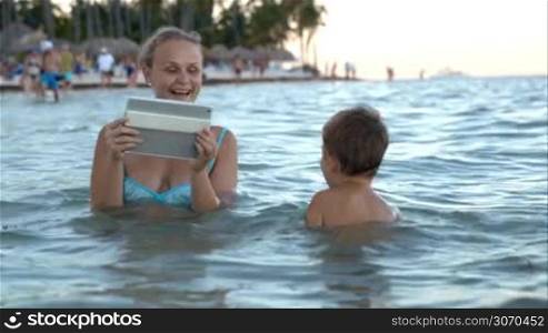 Mother using pad to take picture or make video of little son playing in the sea, then the looking on pad together