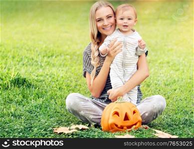 Mother with baby enjoying Halloween holiday outdoors, sitting on fresh green grass field, traditional festive decoration, family fun