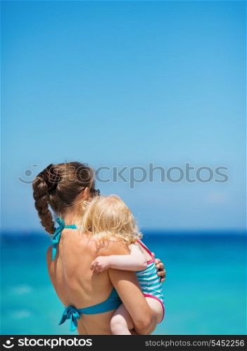 Mother with baby on sea shore looking into distance. Rear view