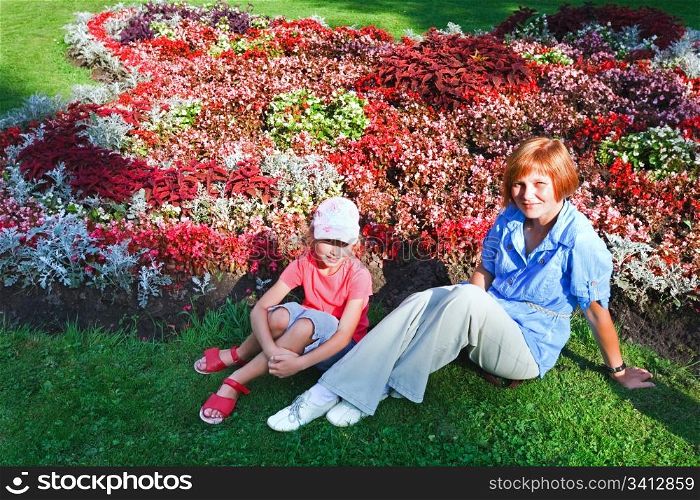 Mother with daughter resting near blossoming colorful flowerbed in summer city park