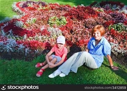 Mother with daughter resting near blossoming colorful flowerbed in summer city park