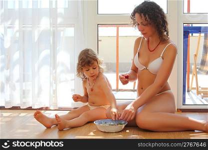 Mother with daughter sit on floor in room and eat cherry
