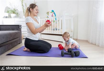 Mother with her baby son sitting on fitness mat at home and exercising with dumbbells