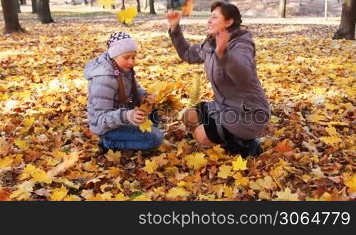 mother with her daughter sit squatted and scatter yellow leaves in beautiful autumn city park