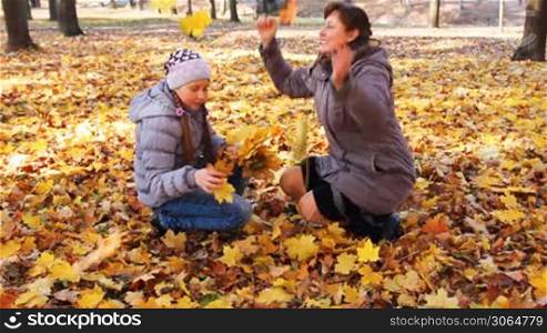 mother with her daughter sit squatted and scatter yellow leaves in beautiful autumn city park