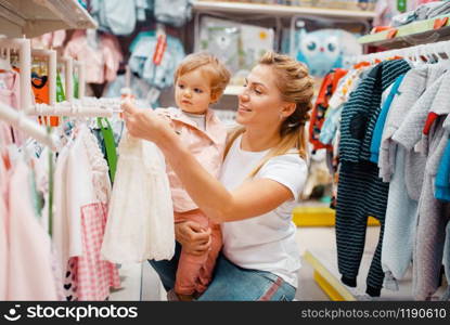 Mother with her little girl choosing clothes in kids store. Mom and child buying dress in supermarket together, family shopping. Mother with girl choosing clothes in kids store