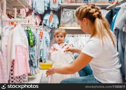 Mother with her little girl choosing clothes in kids store. Mom and child buying dress in supermarket together, family shopping