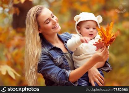 Mother with son enjoying autumn, portrait of a young mom looking at her son, baby explores dry tree leaves, life and weather change concept