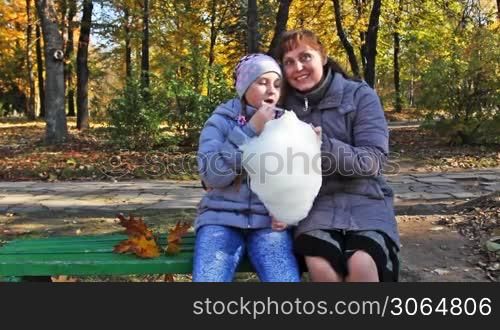 mother with teen daughter sit on bench and eat cotton candy in beautiful autumn city park