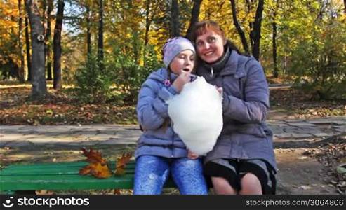 mother with teen daughter sit on bench and eat cotton candy in beautiful autumn city park