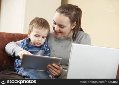 Mother Working On Laptop Whilst Son Uses Digital Tablet