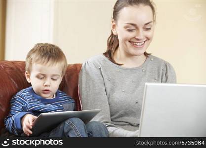 Mother Working On Laptop Whilst Son Uses Digital Tablet