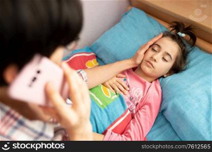 Mother worried about her daughter&rsquo;s temperature calling the doctor. Little girl on bed.. Mother worried about her daughter&rsquo;s temperature calling the doctor