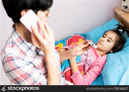 Mother worried about her daughter&rsquo;s temperature calling the doctor. Little girl on bed.. Mother worried about her daughter&rsquo;s temperature calling the doctor