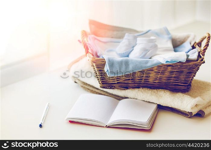 motherhood, care, parenthood and object concept - close up of pile of baby clothes with towel for newborn boy in basket and blank notebook or diary on table