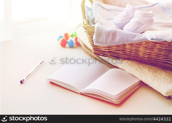 motherhood, care, parenthood and object concept - close up of pile of baby clothes with towel for newborn boy in basket and blank notebook or diary on table