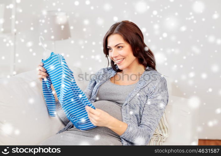 motherhood, pregnancy, people and kids clothing concept - happy woman holding and looking at blue baby boys bodysuit at home over snow
