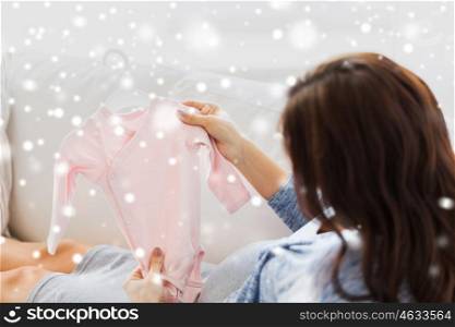 motherhood, pregnancy, people, winter and kids clothing concept - close up of happy woman holding and looking at pink baby girls bodysuit at home over snow