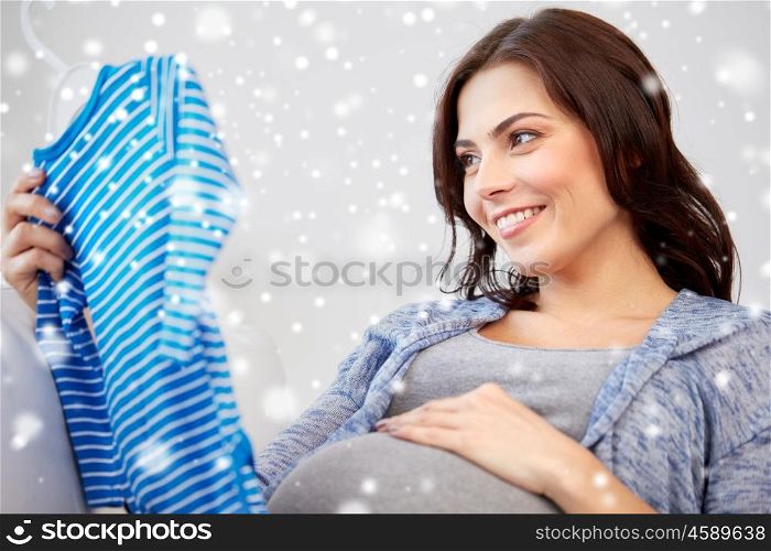motherhood, pregnancy, people, winter and kids clothing concept - happy woman holding and looking at blue baby boys bodysuit at home over snow