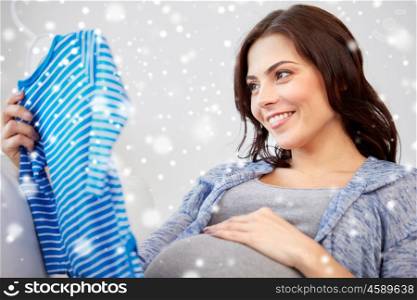 motherhood, pregnancy, people, winter and kids clothing concept - happy woman holding and looking at blue baby boys bodysuit at home over snow