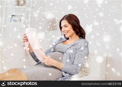 motherhood, pregnancy, people, winter and kids clothing concept - happy woman holding and looking at pink baby girls bodysuit at home over snow
