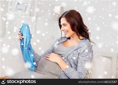 motherhood, pregnancy, winter, people and kids clothing concept - happy woman holding and looking at blue baby boys bodysuit at home over snow