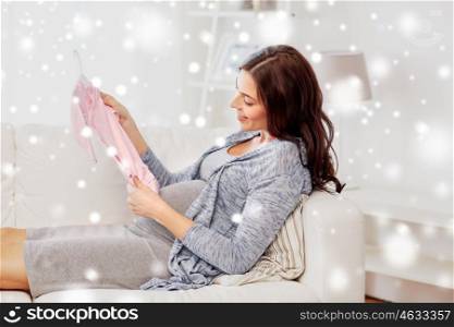 motherhood, pregnancy, winter, people and kids clothing concept - happy woman holding and looking at pink baby girls bodysuit at home over snow