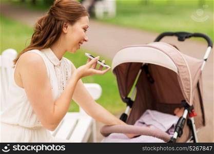 motherhood, technology and people concept - happy mother with baby girl in stroller using voice command recorder on smartphone at summer park. happy mother with smartphone and stroller at park