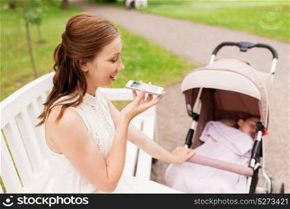 motherhood, technology and people concept - happy mother with baby girl in stroller using voice command recorder on smartphone at summer park. mother with stroller and smartphone at summer park