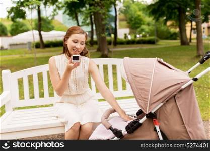 motherhood, technology and people concept - happy mother with baby in stroller using voice command recorder on smartphone at summer park. mother with stroller and smartphone at summer park