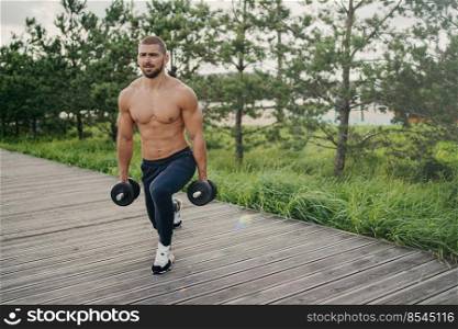 Motivated bearded man with naked torso makes squat with heavy barbells, does physical exercises outdoor, enjoys workout, poses near green trees, doing weightlifting. Healthy lifestyle concept