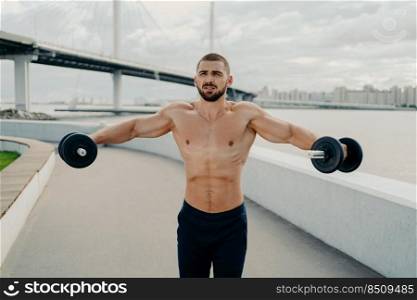 Motivated bodybuilder raises barbells outdoor enjoys great training trains muscles concentrated somewhere poses with naked muscular torso. Athletic shirtless sportsman has workout in open air