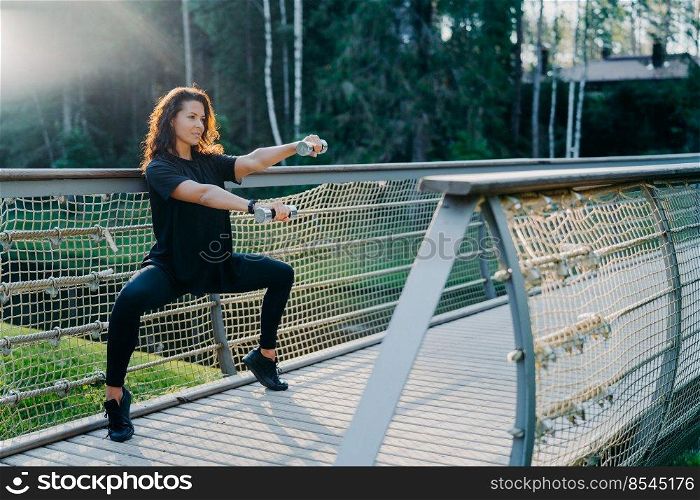 Motivated brunette active young woman does squat exercises with dumbbells, trains biceps, dressed in black active wear, poses at bridge outdoor during sunrise, has morning workout. Sport concept