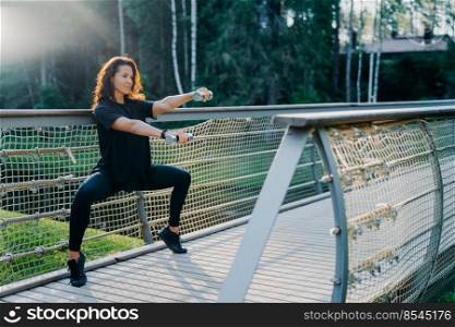 Motivated brunette active young woman does squat exercises with dumbbells, trains biceps, dressed in black active wear, poses at bridge outdoor during sunrise, has morning workout. Sport concept