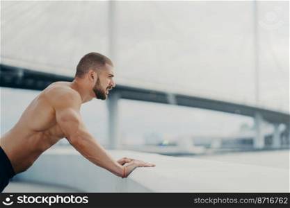 Motivated male sportsman does push up exercise poses outdoor at bridge concentrated into distance has morning workout naked muscular torso warms up before jogging. Healthy lifestyle concept.