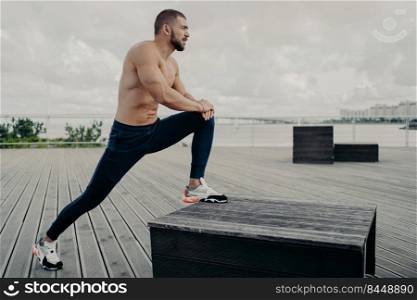 Motivated sportsman leans at knee and does sport exercises outdoor, warms up before jogging, poses near river and dressed in active wear. Male runner does stetching exercise, prepares for workout