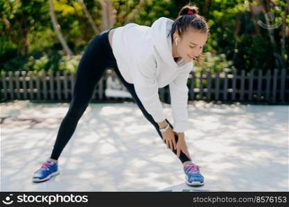 Motivated young adult woman goes in for sport regularly reaches hand to foot leans down has feet shoulder width apart wears sportsclothes enjoys training outdoor sports activity for weight loss