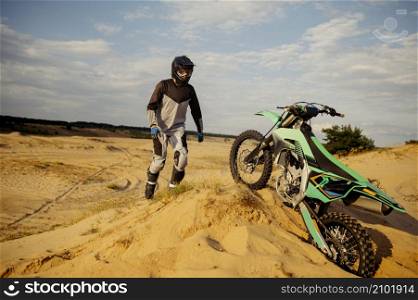 Motocross rider walking to motorbike trapped in sand. Driver and speed motorcycle. Extreme sport. Driver walking to motorbike trapped in sand
