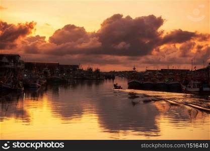 motor fishing boat sailing through large cargo boats at Pak Nam river port or dock, Rayong Thailand. Transportation at sunrise with motion water and light reflection with twilight sky.