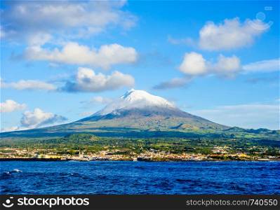Mount Pico volcano western slope and town of Madalena viewed from ocean under blue sky and clouds, in Azores, Portugal.