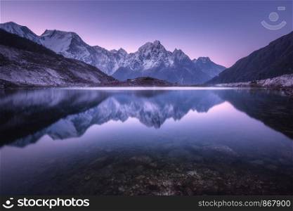 Mountain lake with perfect reflection at sunrise. Beautiful landscape with purple sky, snowy mountains, hills, fog over the lake at twilight in Nepal. Snow covered rocks is reflected in water. Nature