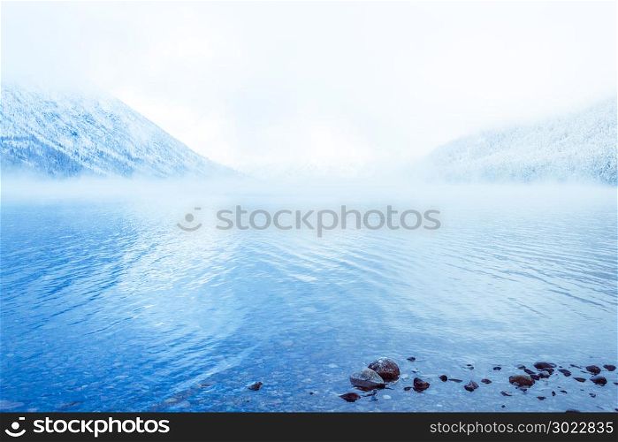 Mountain lake with rocks on the shore, winter fog over the water surface. Travel to the mountains on foot, wildlife.
