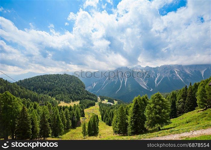 Mountain panorama with forest, Dolomites, valley and chairlift in summer. Sky with clouds.