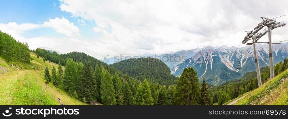 Mountain panorama with forest, Dolomites, valley and chairlift in summer. Sky with clouds.