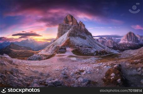 Mountain pass and beautiful sky with colorful clouds at sunset. Amazing panoramic landscape with rocks, mountain peaks, stones, trails, buildings, trees on hills at dusk. Autumn in Dolomites, Italy