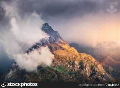Mountain peak in low clouds at colorful sunset in Nepal. Dramatic landscape with beautiful high rocks, dramatic sky, sunlight, tress, orange grass in fog at sunset. Nature. Himalayan mountains. Travel