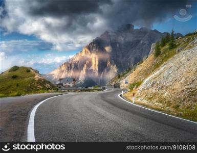 Mountain road at sunny day in summer. Dolomites, Italy. Beautiful roadway, green tress, high rocks in clouds, blue sky. Landscape with empty highway through the mountain pass in spring. Travel